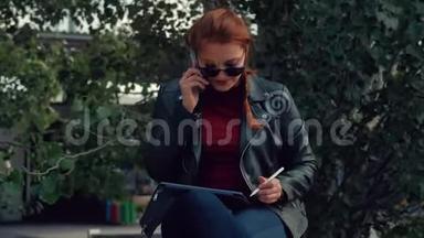 戴墨镜的年轻女子坐在公园里的长凳上，一边用手机说话，一边记录着在公园里的谈话